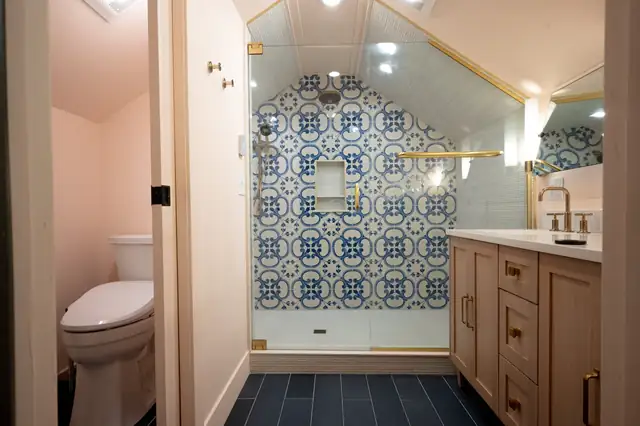 Bathroom remodel with a blue and white patterned tile shower under a sloped ceiling, gold fixtures, wood vanity and dark floor tile