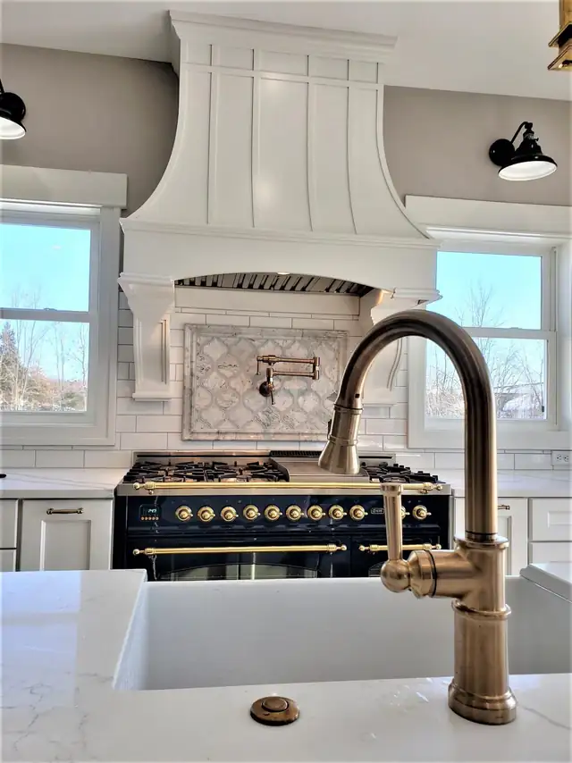 White kitchen remodel with a black and brass gas range, curved white hood, marble counters and a brass faucet over a farmhouse sink